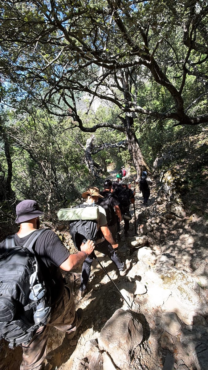 A hiker with a backpack following the group up a shaded rocky trail through trees